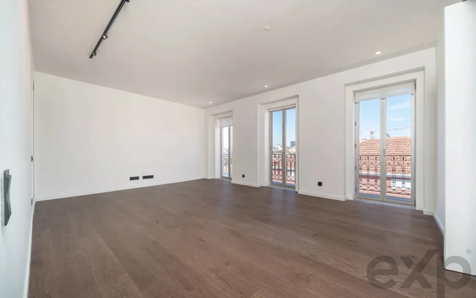 Sala ampla de apartamento remodelado no Rato, Lisboa, com soalho de madeira e três janelas de sacada sobre os telhados